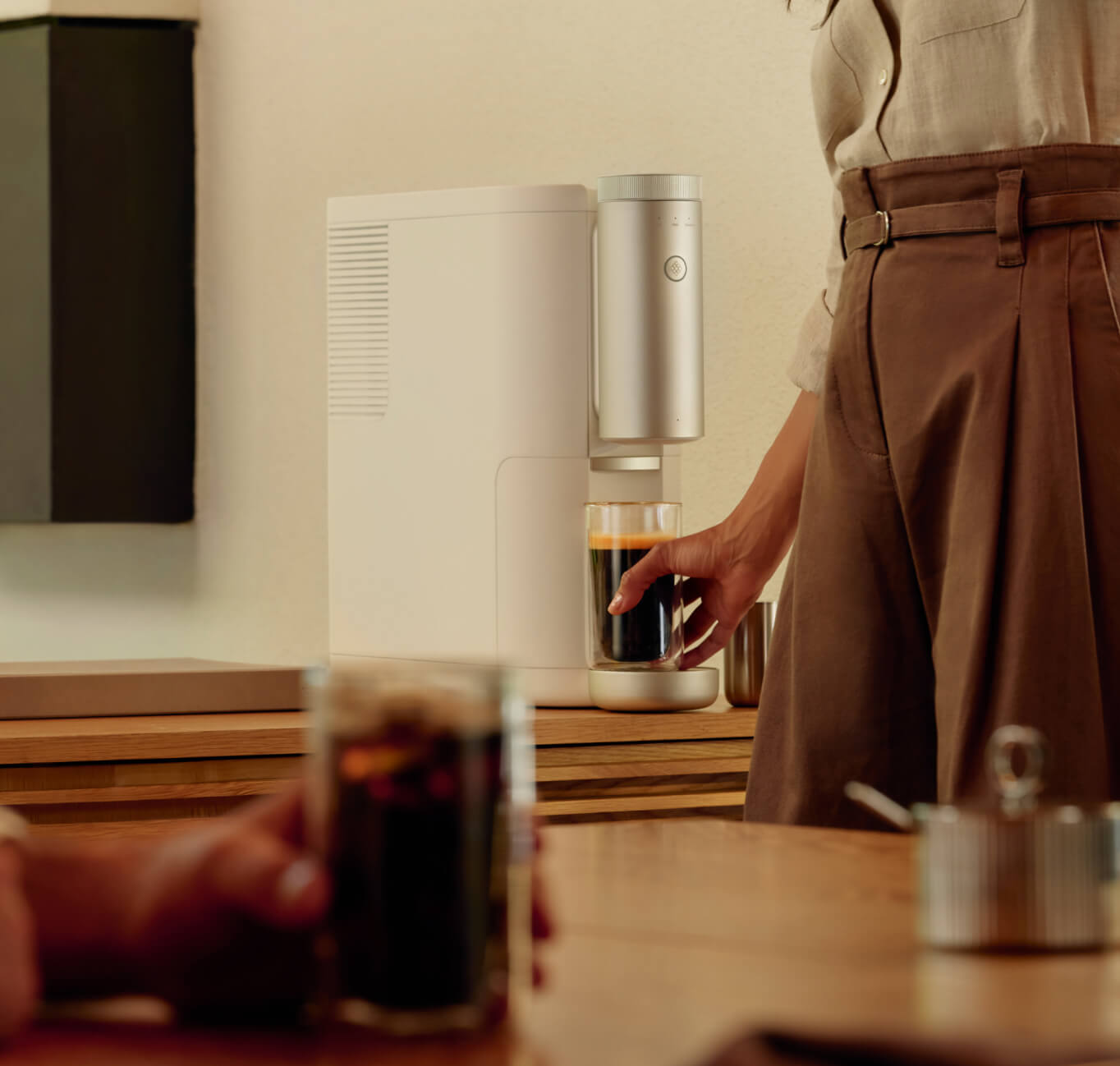 A hand holding a cup of coffee resting on a table in the foreground and a person grabbing a finished cup of coffee from a white Cumulus Machine in the background