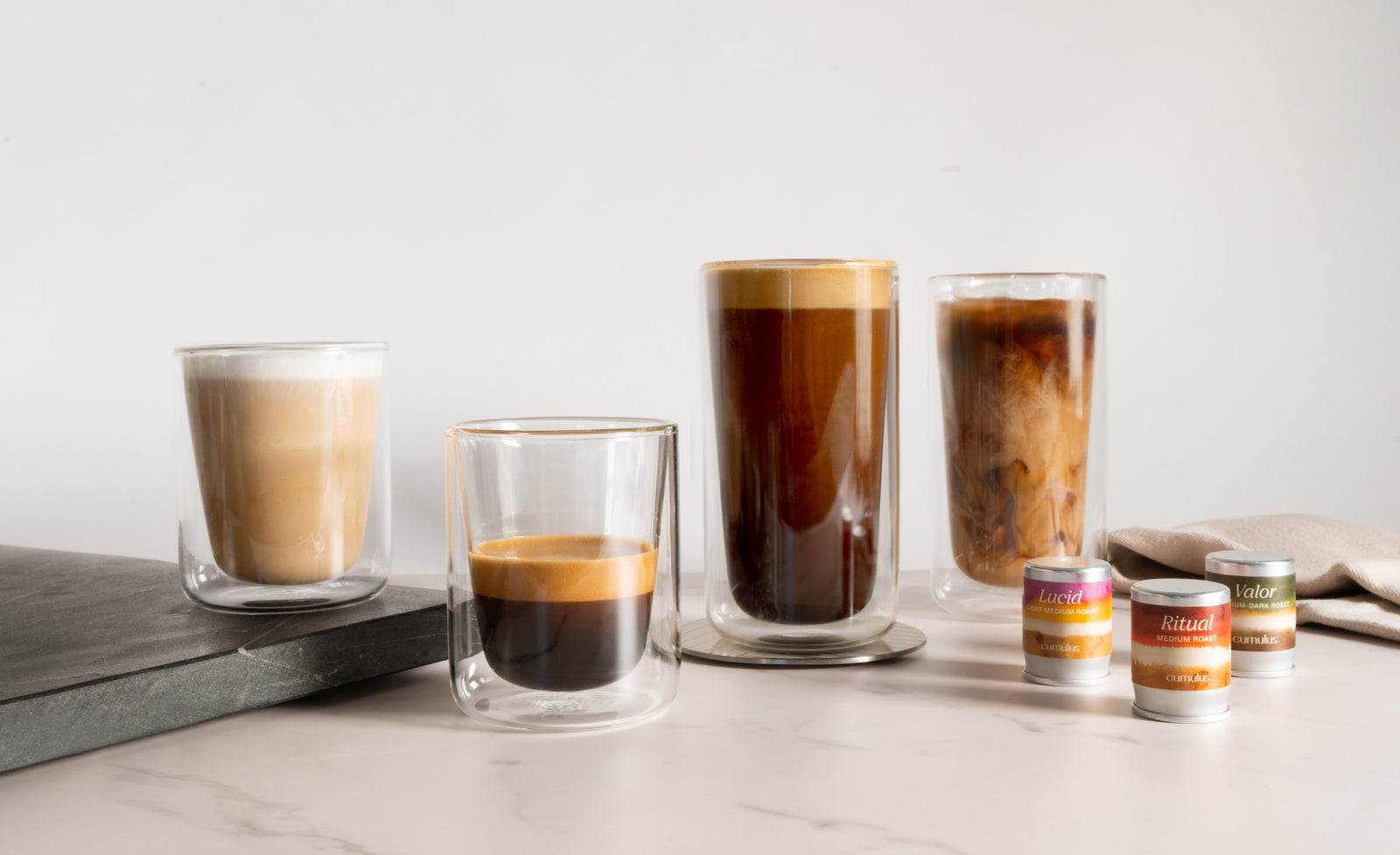 2 short glasses and 2 tall glasses filled with different kinds of Cumulus cold brew coffee. Next to them on the counter are 3 capsules: Ritual, Lucid, and Valor.