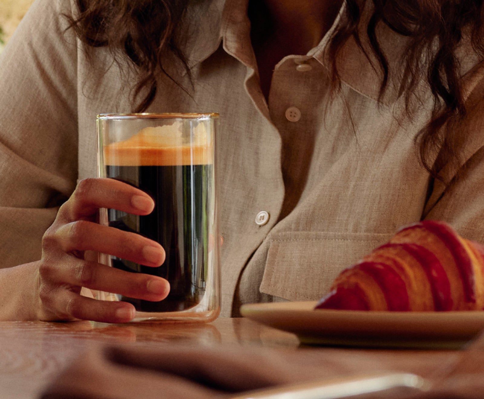 A zoomed shot of a woman holding a cup of coffee next to a croissant