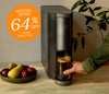 A sleek black coffee maker on a counter next to a plant and bowl of apples + pears. A badge says "Special offer - 64% off limited quantities"