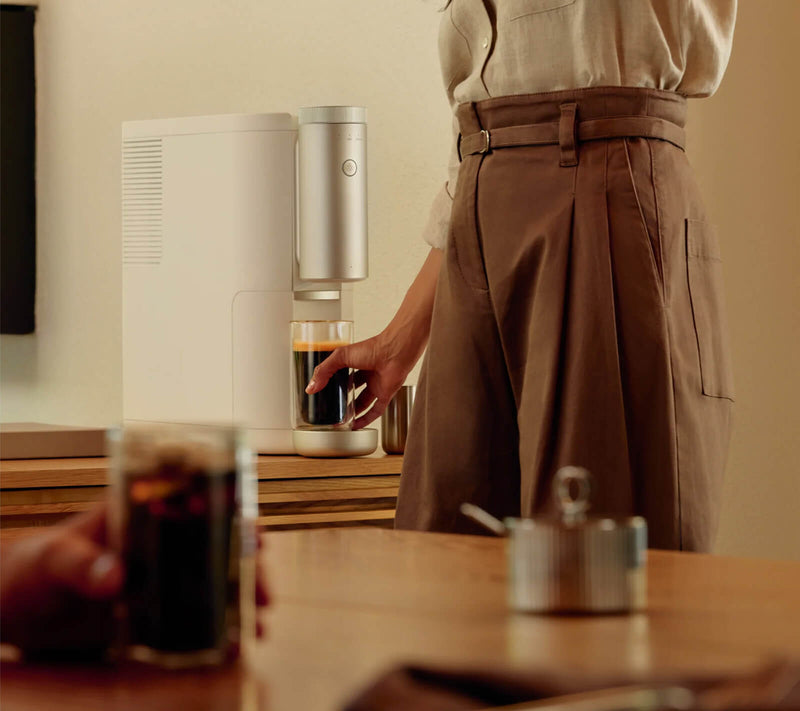 Person in foreground holding a cup of coffee, person in background retrieving a completed cup of coffee from a Cumulus machine
