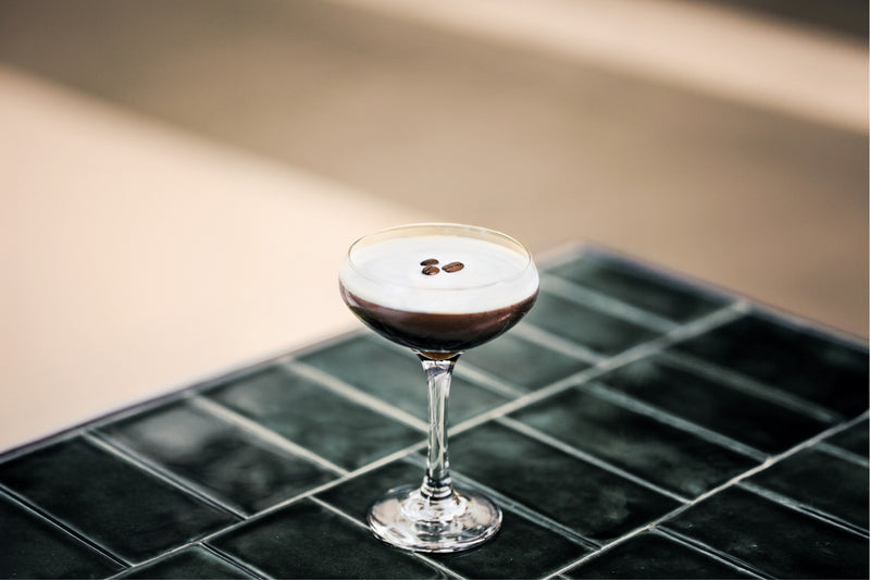 An espresso martini sitting on a tile countertop with 3 coffee beans floating in the middle