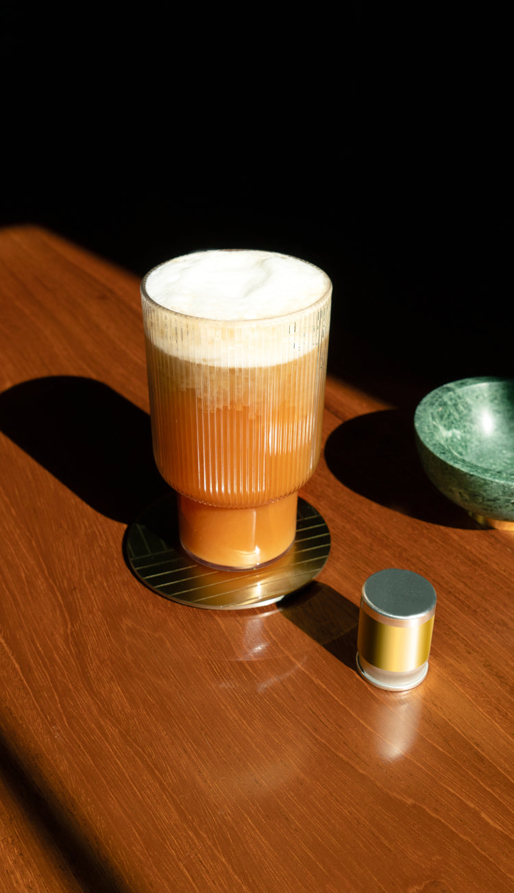 A glass of coffee with a thick head of foam on it sitting on a wooden table next to an unlabeled cumulus capsule.
